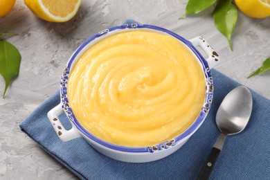 Delicious lemon curd in bowl, fresh citrus fruit, green leaves and spoon on grey table