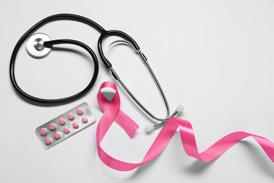 Breast cancer awareness. Pink ribbon, stethoscope and pills on white background, flat lay. Space for text