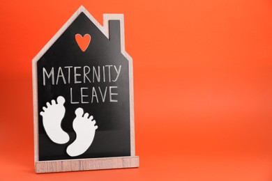 Photo of Wooden house figure with words Maternity Leave and paper cutout of baby feet on orange background. Space for text