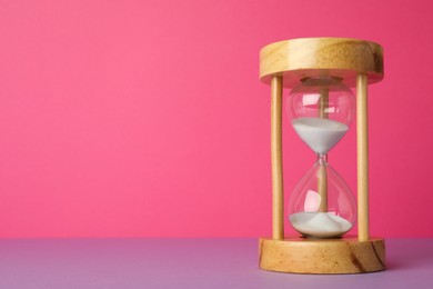 Photo of Hourglass with flowing sand on violet table against pink background, space for text. Menopause concept
