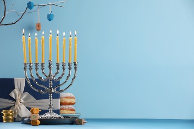 Photo of Hanukkah celebration. Menorah with burning candles, dreidels, donuts and gift box on table against light blue background. Space for text