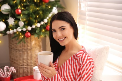Photo of Young woman with cup of coffee near Christmas tree at home