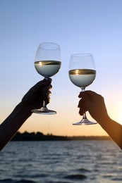 Photo of Women clinking glasses of wine near river at sunset, closeup