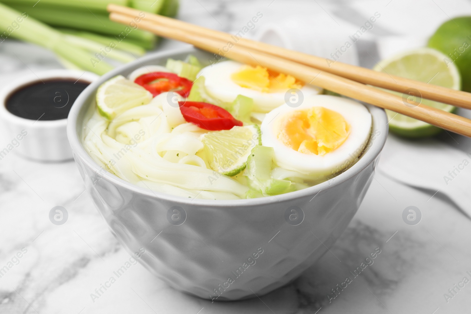 Photo of Bowl of delicious rice noodle soup with celery and egg on white marble table, closeup