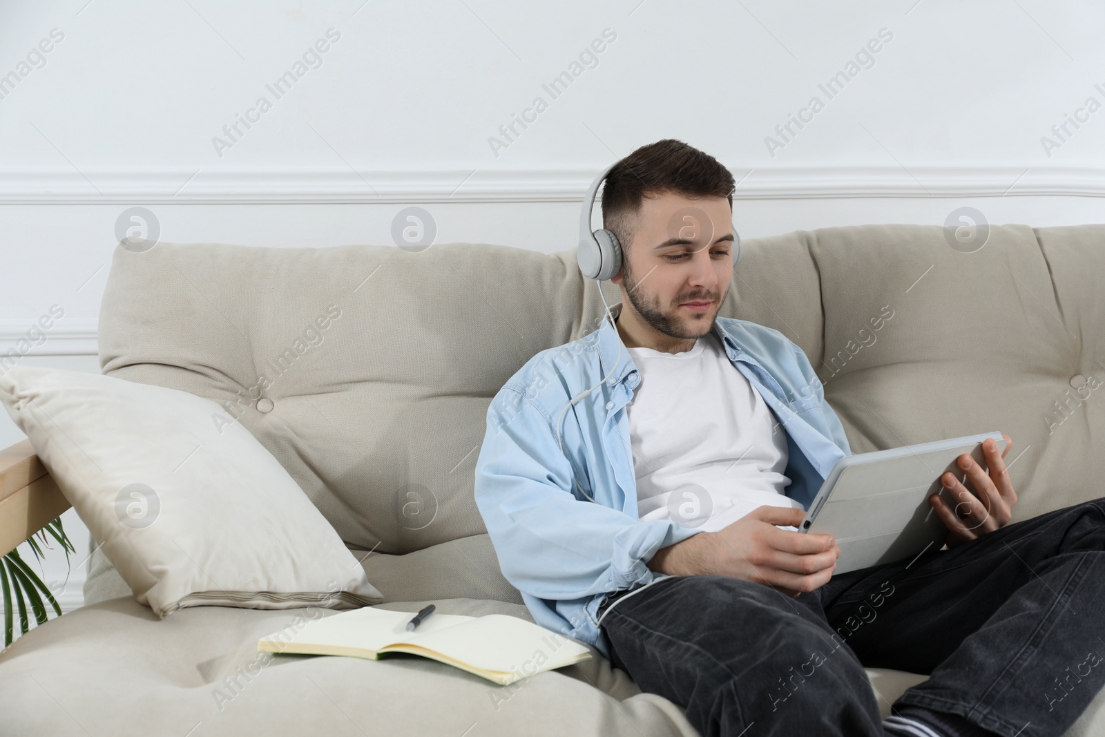 Photo of Young man with headphones using modern tablet for studying on sofa at home. Distance learning