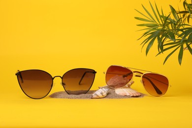 Photo of Stylish sunglasses, seashells, sand and palm leaves on yellow background