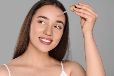 Photo of Beautiful young woman applying essential oil onto face on grey background