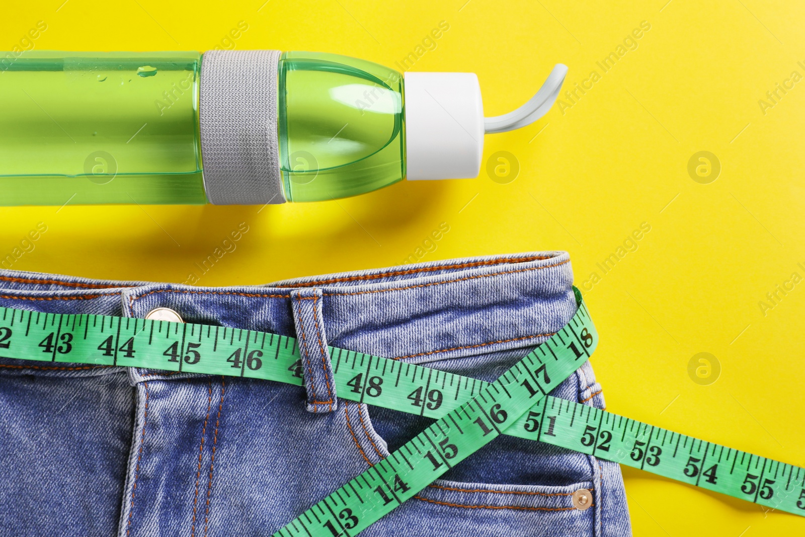 Photo of Jeans with measuring tape and bottle of water on yellow background, flat lay. Weight loss concept