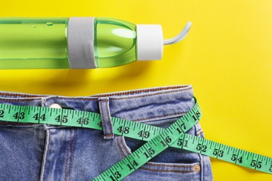 Photo of Jeans with measuring tape and bottle of water on yellow background, flat lay. Weight loss concept