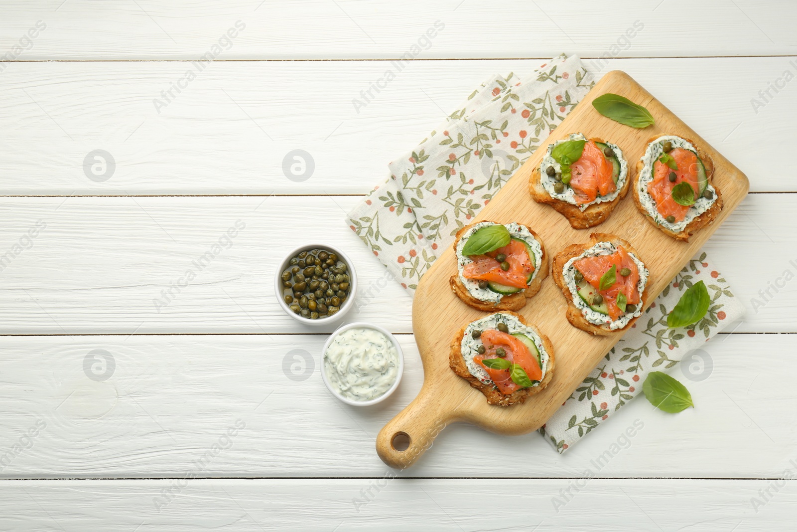 Photo of Tasty canapes with salmon, capers, cucumber and sauce on white wooden table, top view. Space for text