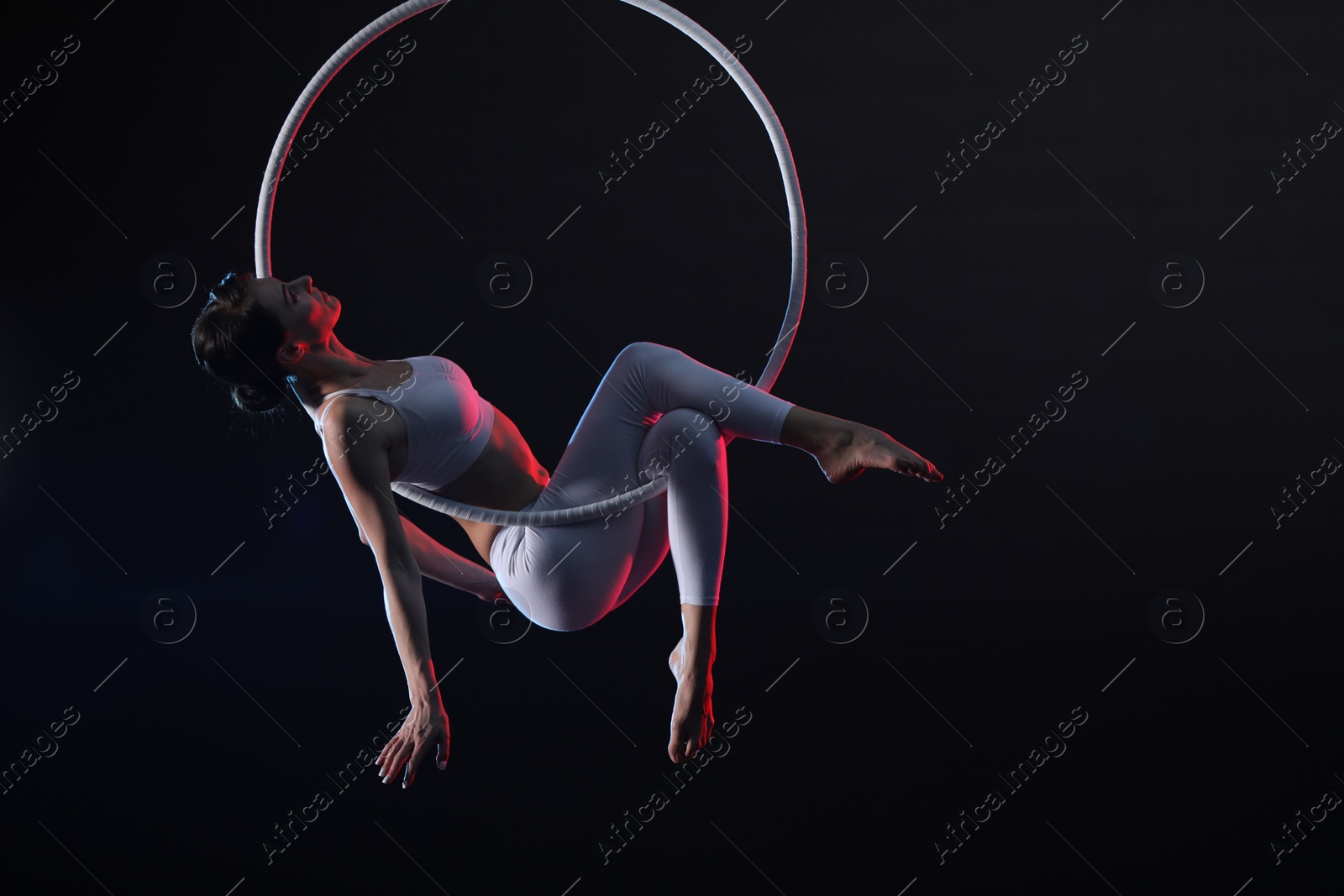 Photo of Young woman performing acrobatic element on aerial ring against dark background