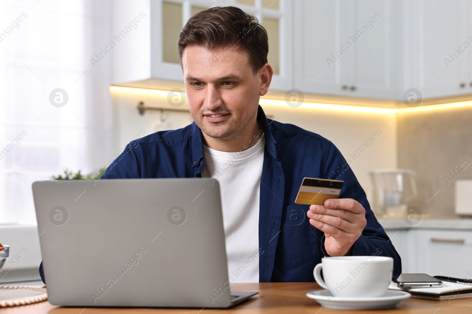 Photo of Man with credit card and laptop shopping online at wooden table in kitchen