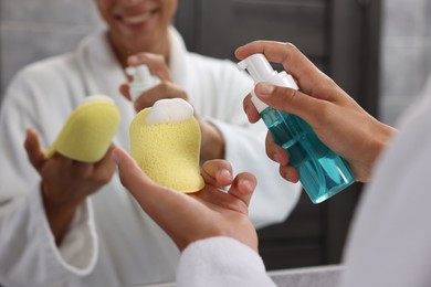 Photo of Happy young man applying face cleanser on sponge near mirror in bathroom, closeup