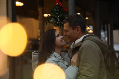 Photo of Happy couple standing under mistletoe bunch outdoors, bokeh effect