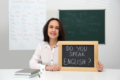 Photo of Teacher holding small chalkboard with inscription Do You Speak English? at table in classroom