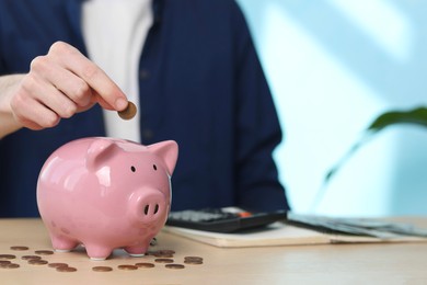 Financial savings. Man putting coin into piggy bank at wooden table, closeup