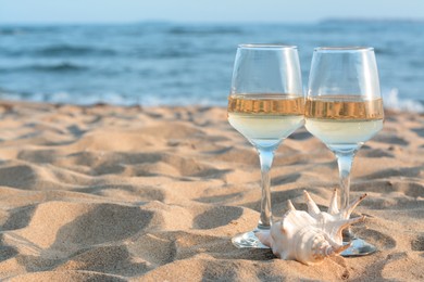 Photo of Glasses of tasty wine and seashell on sand near sea, space for text