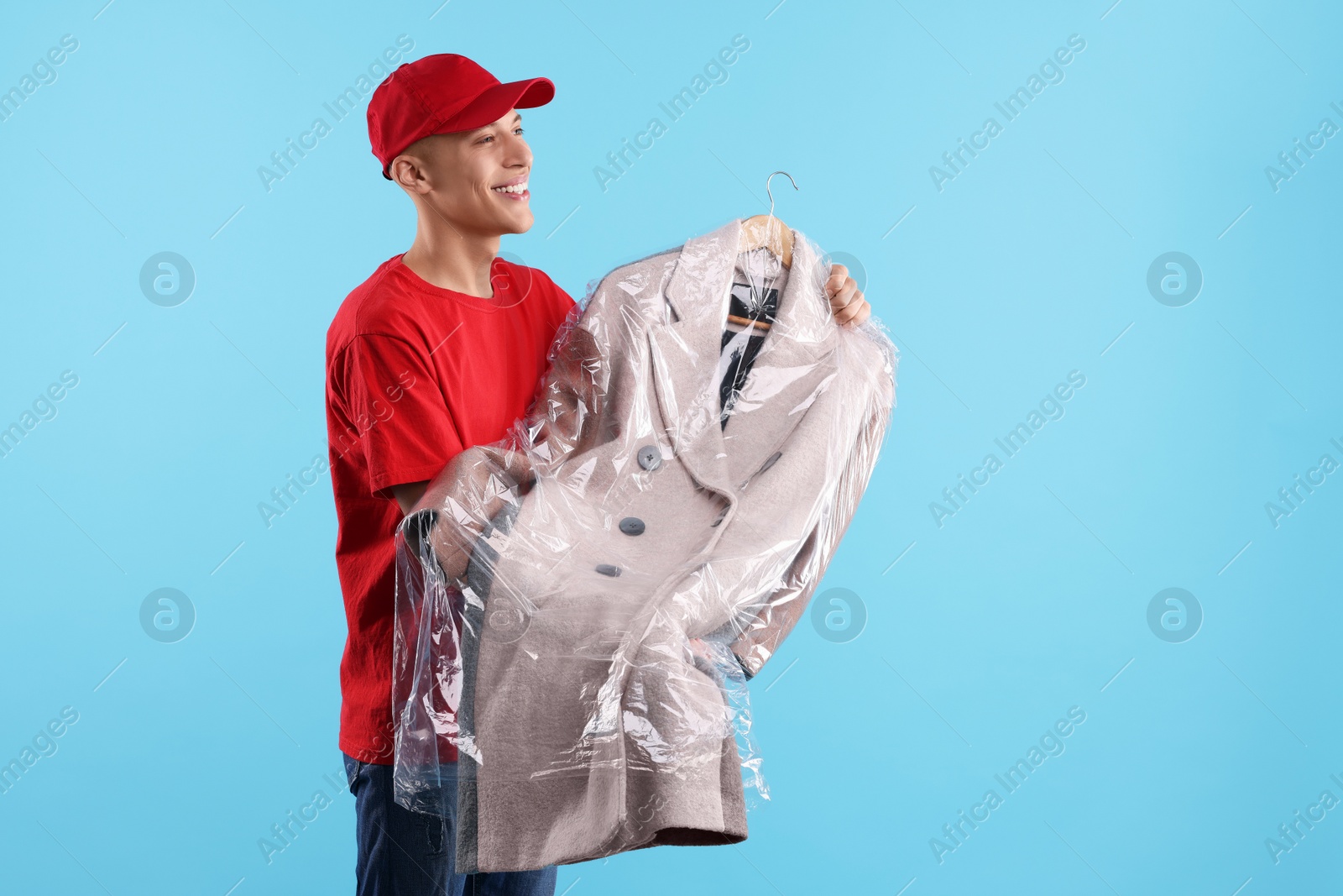 Photo of Dry-cleaning delivery. Happy courier holding coat in plastic bag on light blue background, space for text