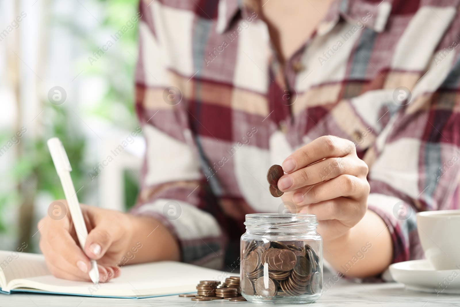 Photo of Woman putting money into glass jar at white marble table, closeup