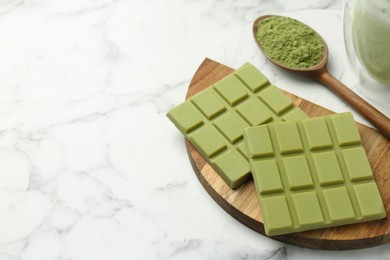 Pieces of tasty matcha chocolate bar and powder in spoon on white marble table, closeup. Space for text