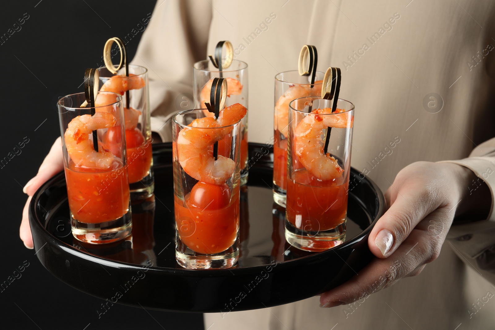 Photo of Woman holding tasty canapes with shrimps, tomatoes and sauce in shot glasses on black background, closeup