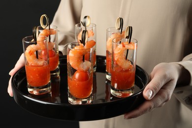 Photo of Woman holding tasty canapes with shrimps, tomatoes and sauce in shot glasses on black background, closeup