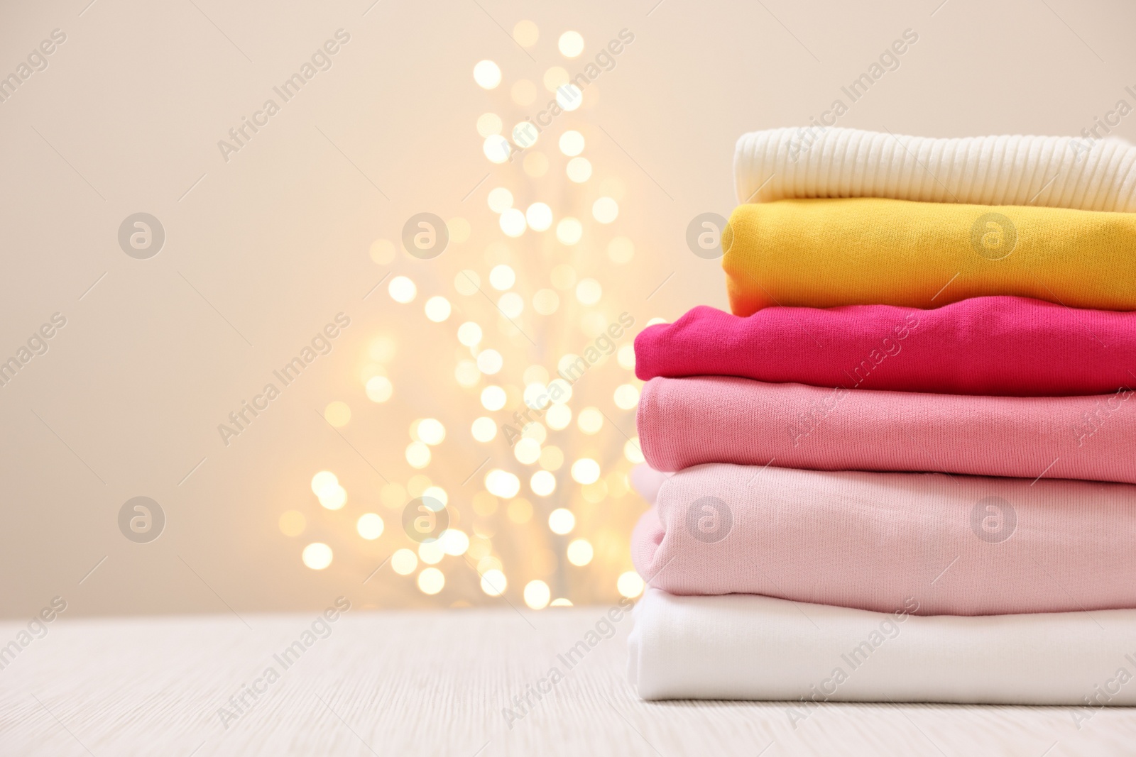 Stack of folded clothes on wooden table against beige background with blurred lights, space for text Photo of Stack of folded clothes on wooden table against beige background with blurred lights, space for text
