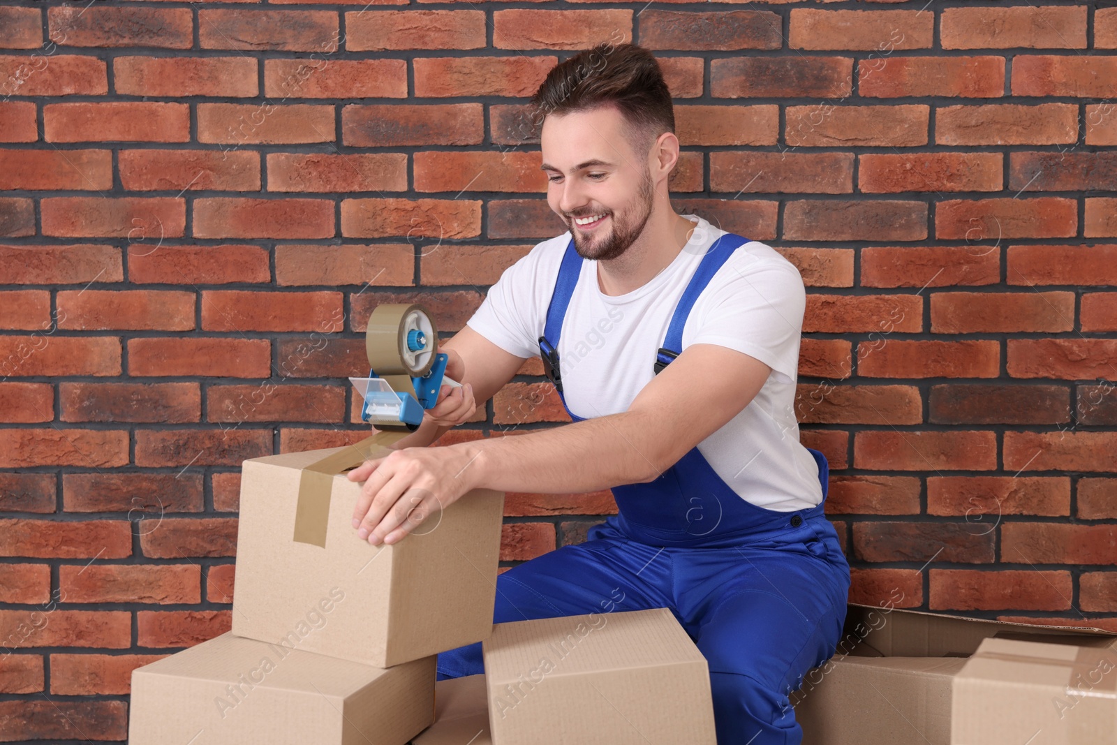 Photo of Smiling worker taping box with adhesive tape dispenser near brick wall