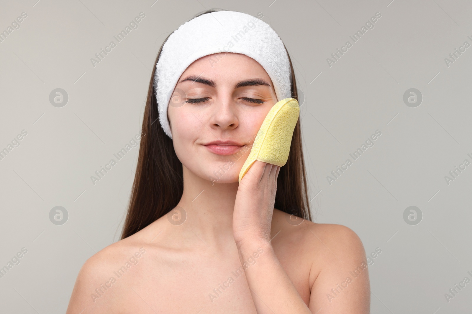 Photo of Young woman with headband washing her face using sponge on light grey background