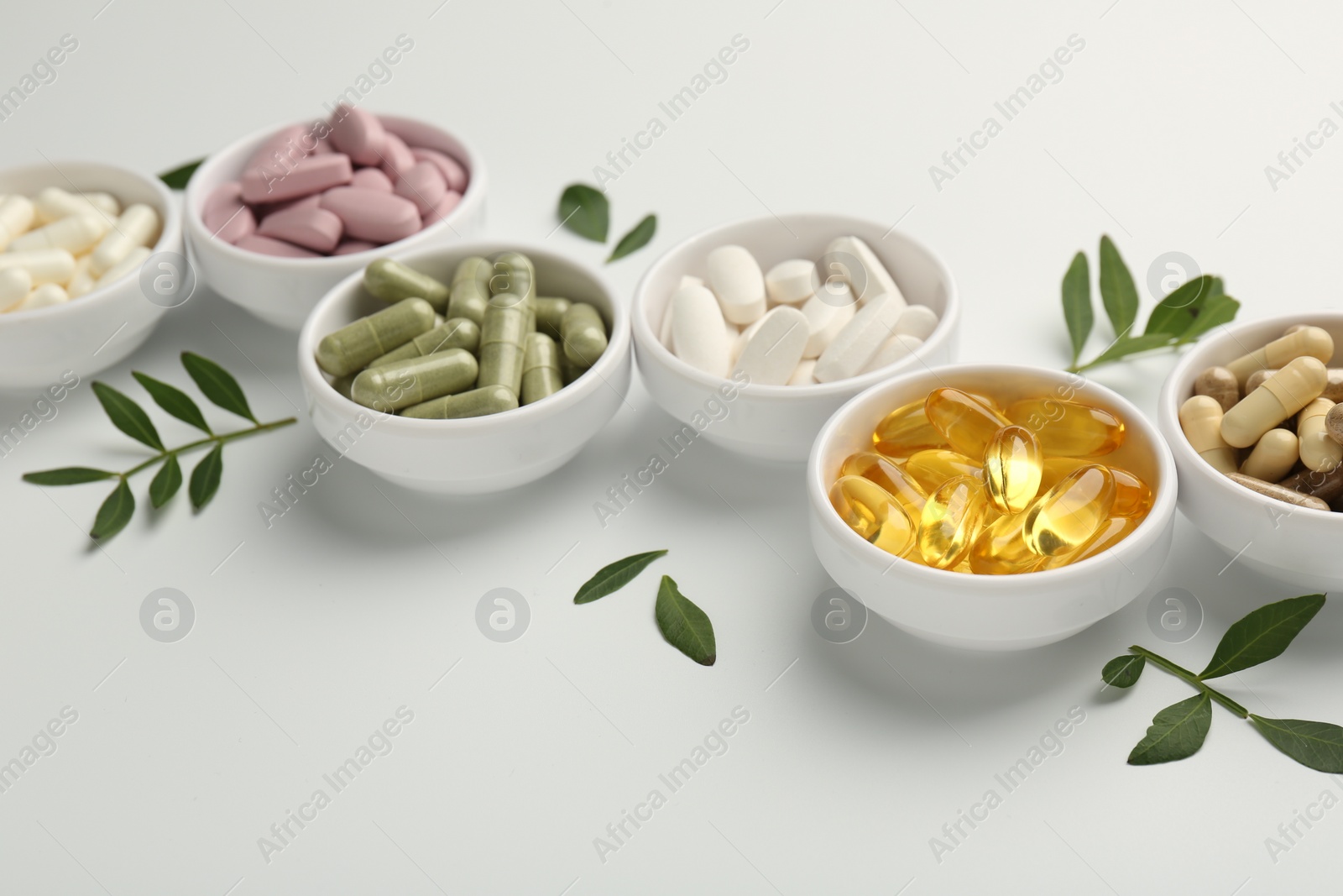 Photo of Different vitamin capsules in bowls and leaves on white background, closeup