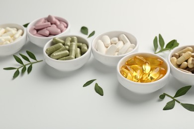 Different vitamin capsules in bowls and leaves on white background, closeup