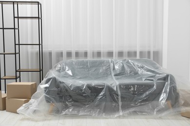 Photo of Stylish sofa covered with plastic film, shelving unit and boxes at home