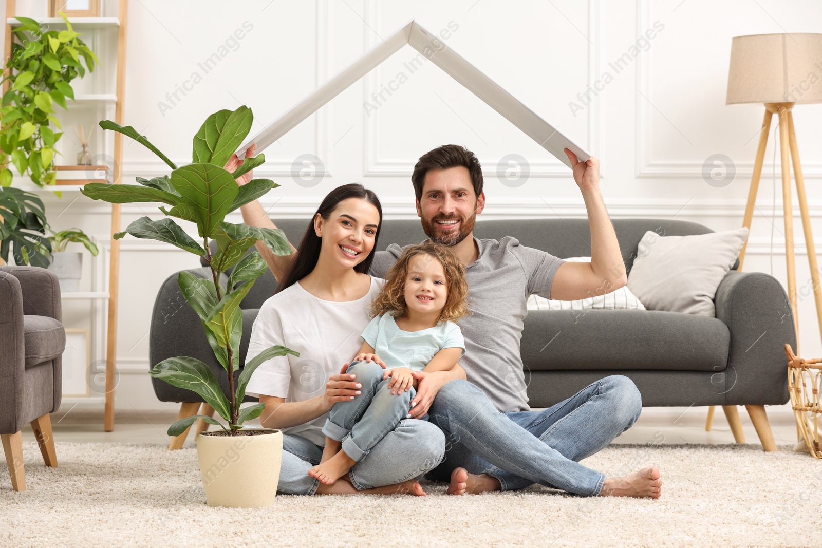 Photo of Housing concept. Happy husband holding plastic roof over his family while sitting on floor at home