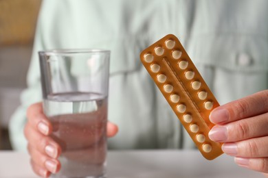 Photo of Woman holding blister of oral contraceptive pills and glass with water at white table, closeup