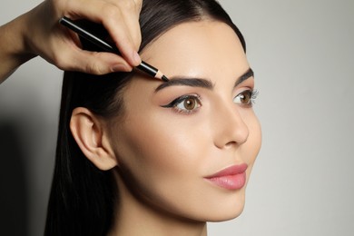 Artist correcting woman's eyebrow shape with pencil on grey background, closeup