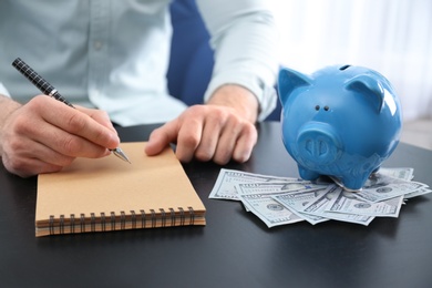 Photo of Businessman with notebook, piggy bank and money at table indoors, closeup