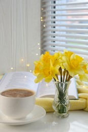 Beautiful yellow daffodils in vase, cup of coffee, book and festive lights on windowsill. Space for text