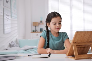 Photo of Little girl doing homework with tablet at table in bedroom