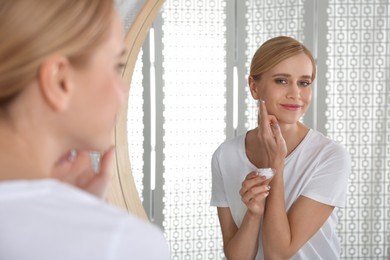 Happy young woman applying cream onto face near mirror in bathroom