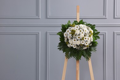 Funeral wreath of flowers on wooden stand near light grey wall, space for text