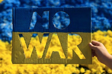 Photo of Woman holding poster in colors of Ukrainian flag with words No War and beautiful blue and yellow flowers on background, closeup