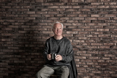 Photo of Poor elderly man holding mug near brick wall