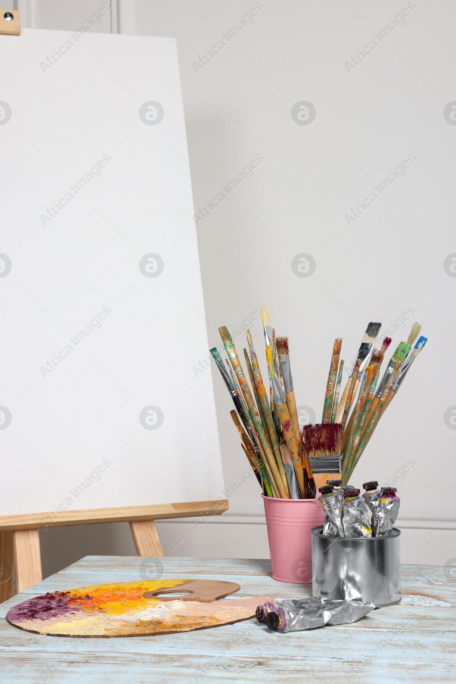 Photo of Easel with blank canvas and different art supplies on wooden table near white wall