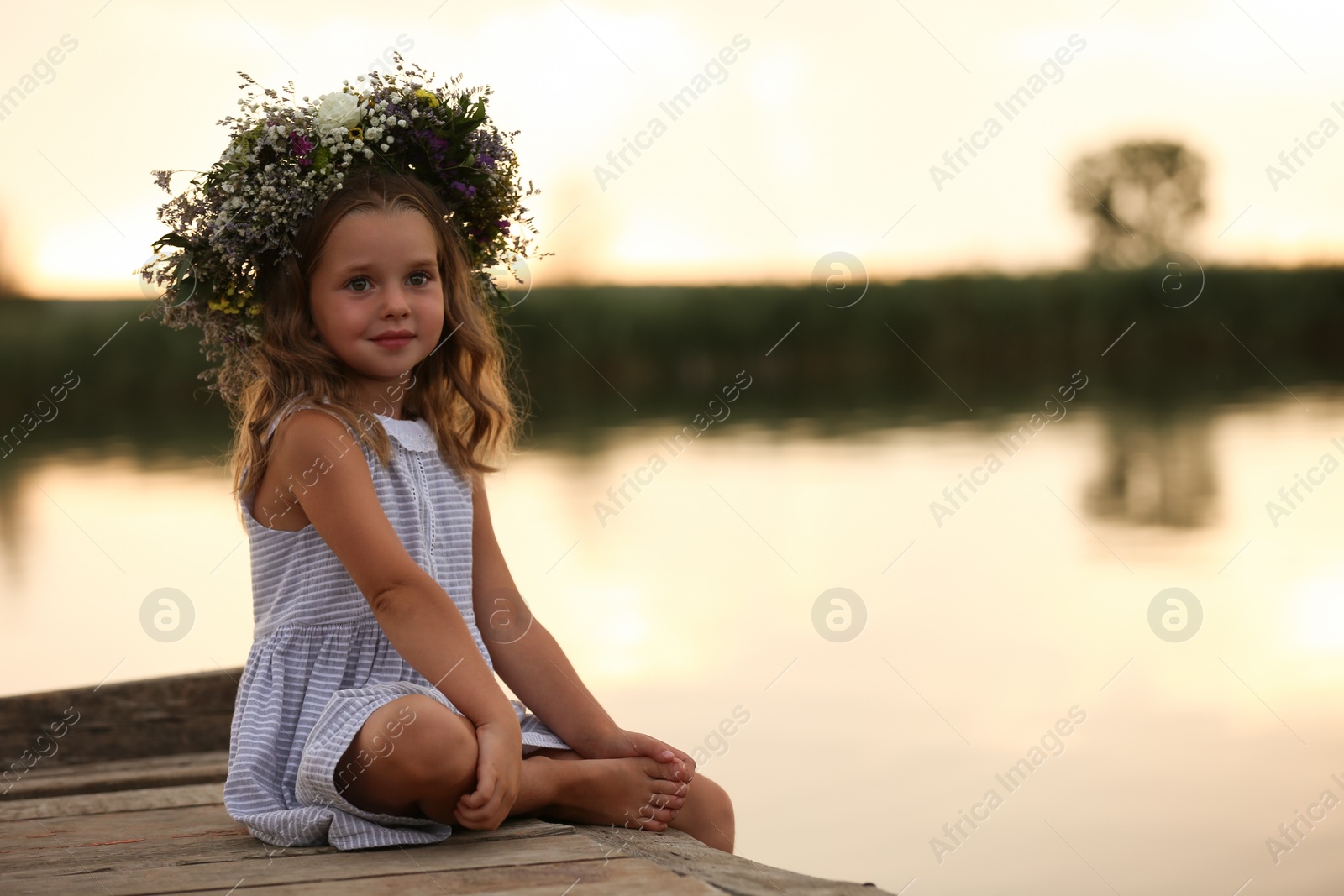 Photo of Cute little girl wearing wreath made of beautiful flowers on pier near river