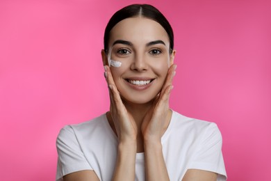 Young woman applying facial cream on pink background