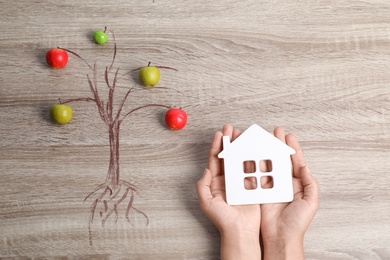 Photo of Woman holding house model near drawing of tree with small apples on wooden background, top view