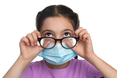 Little girl wiping foggy glasses caused by wearing medical face mask on white background. Protective measure during coronavirus pandemic