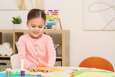 Photo of Cute little girl making colorful paper card at desk in room. Home workplace