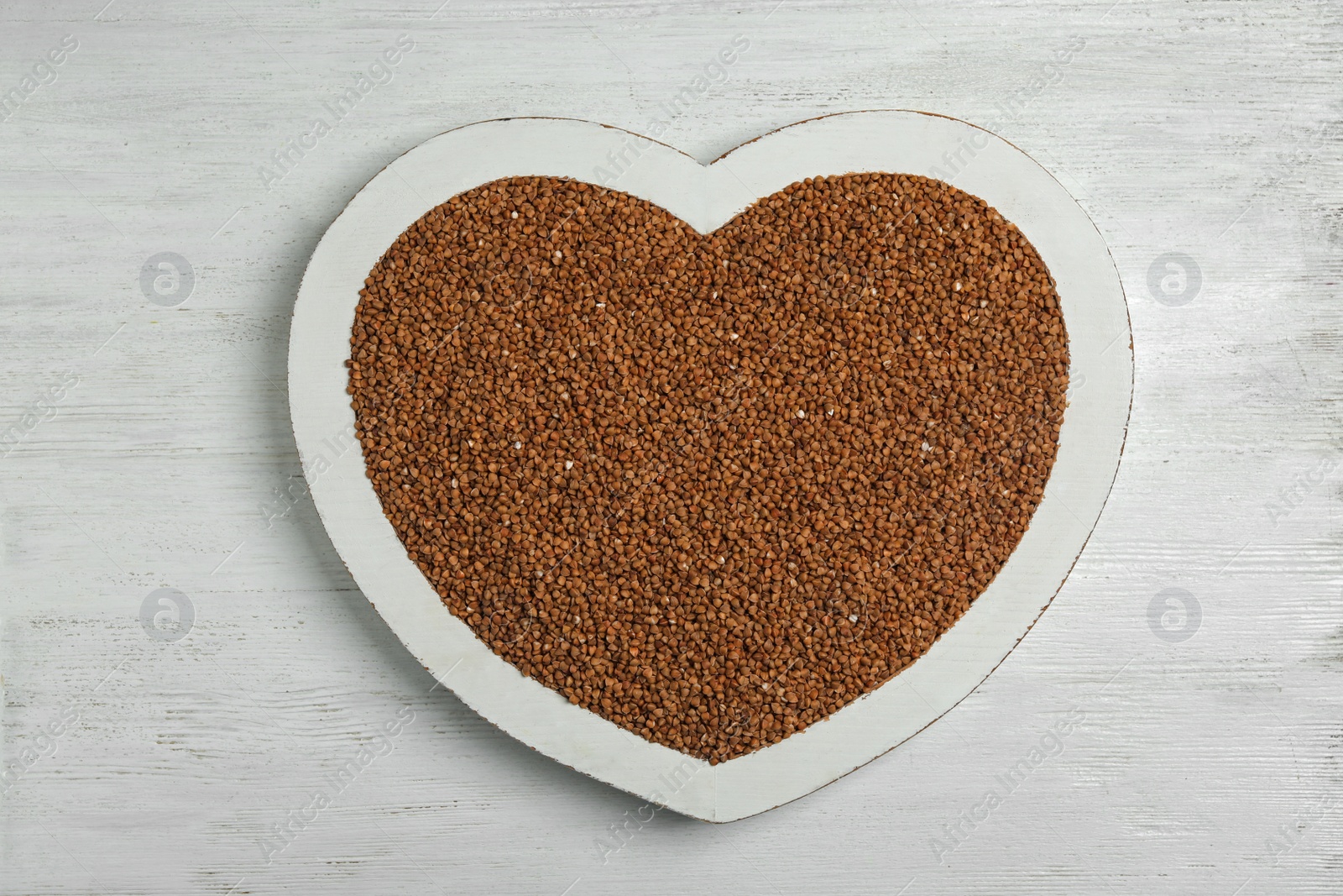 Photo of Heart shaped tray with raw buckwheat on wooden background, top view