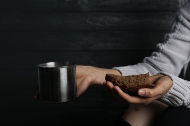 Poor woman holding cup and piece of bread against wooden background, closeup